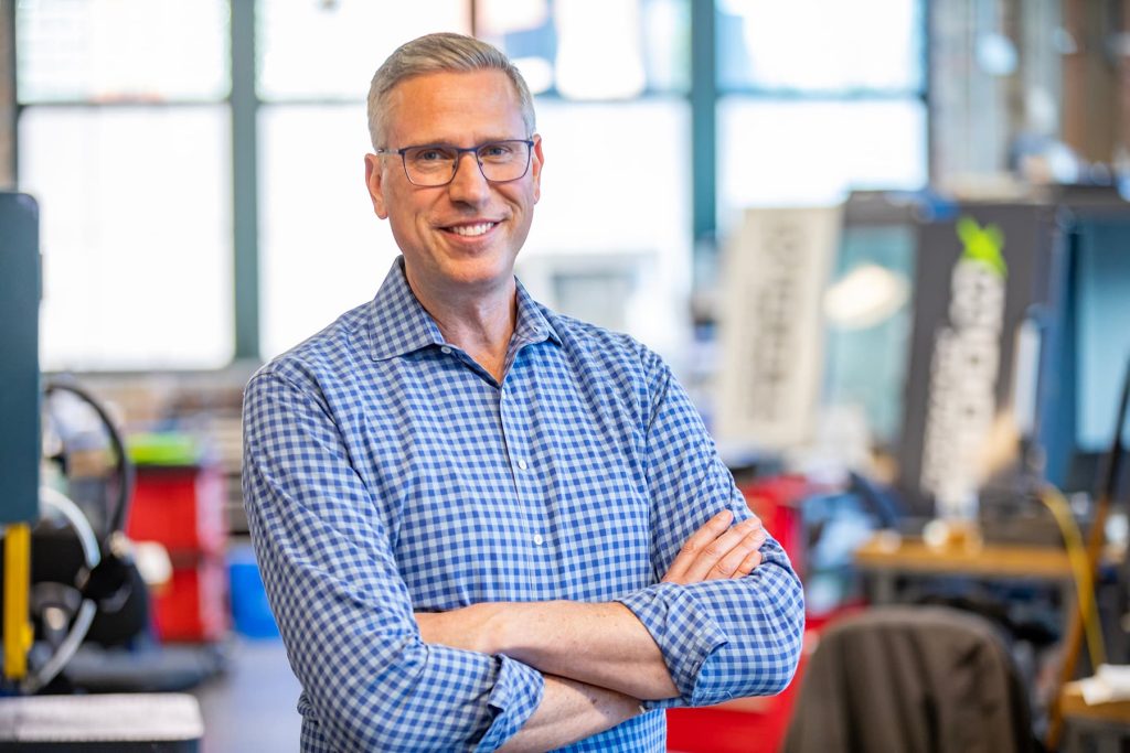 Elected official in a blue checked shirt in a workshop smiles at the camera.