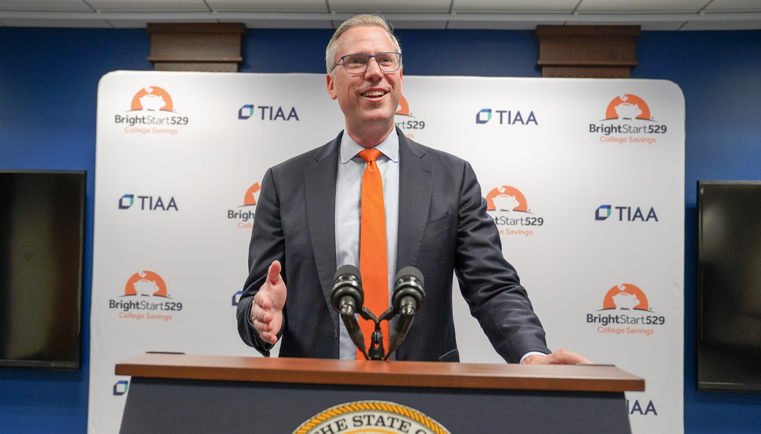 Treasurer Frerichs smiles as he speaks from a podium. Backdrop has the Bright Start logo