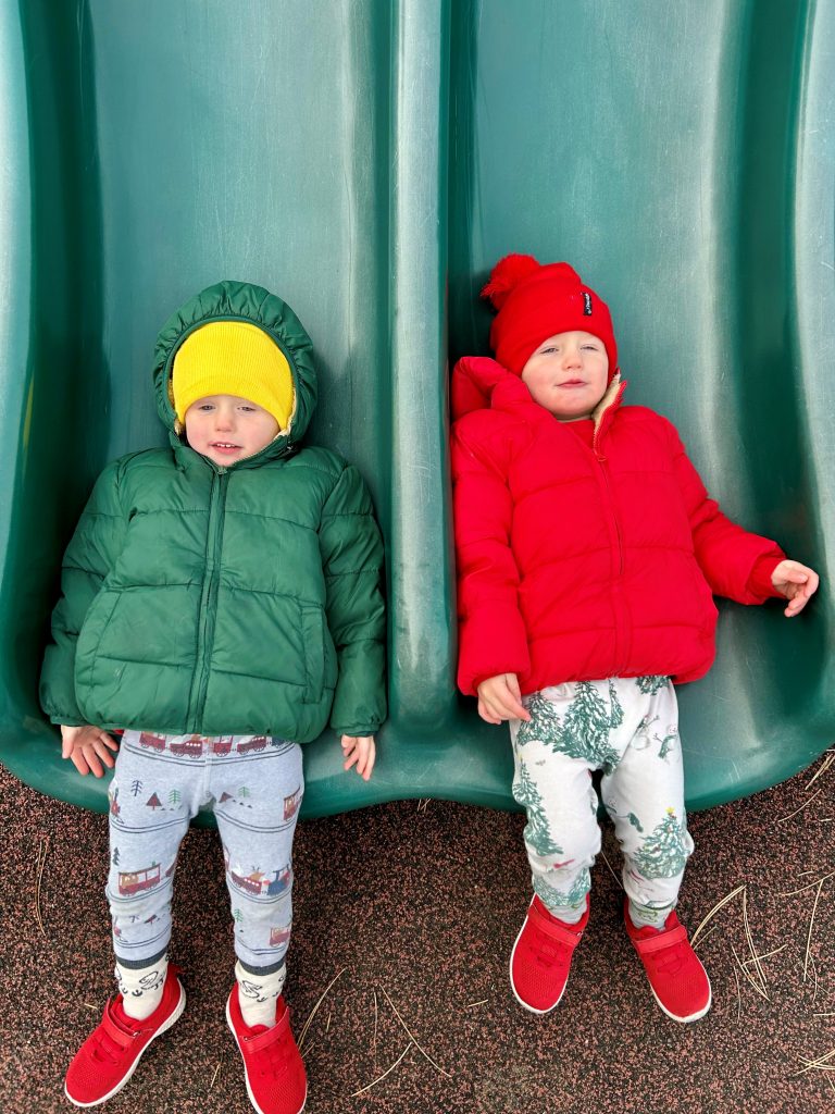 two boys under 3 years old on the bottom of a slide with red and green jackets