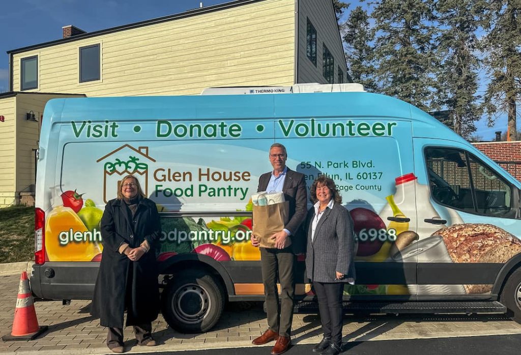 a man holding a bag of groceries stands with two women in front of a food pantry van smiling