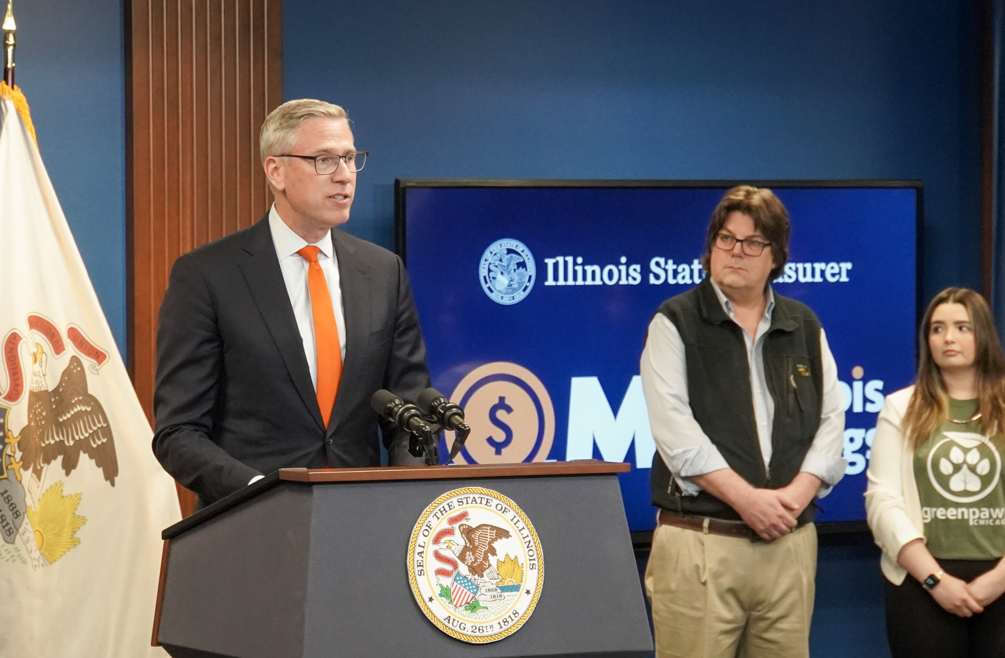 Treasurer Frerichs speaks from a podium as two people stand beside him on his left