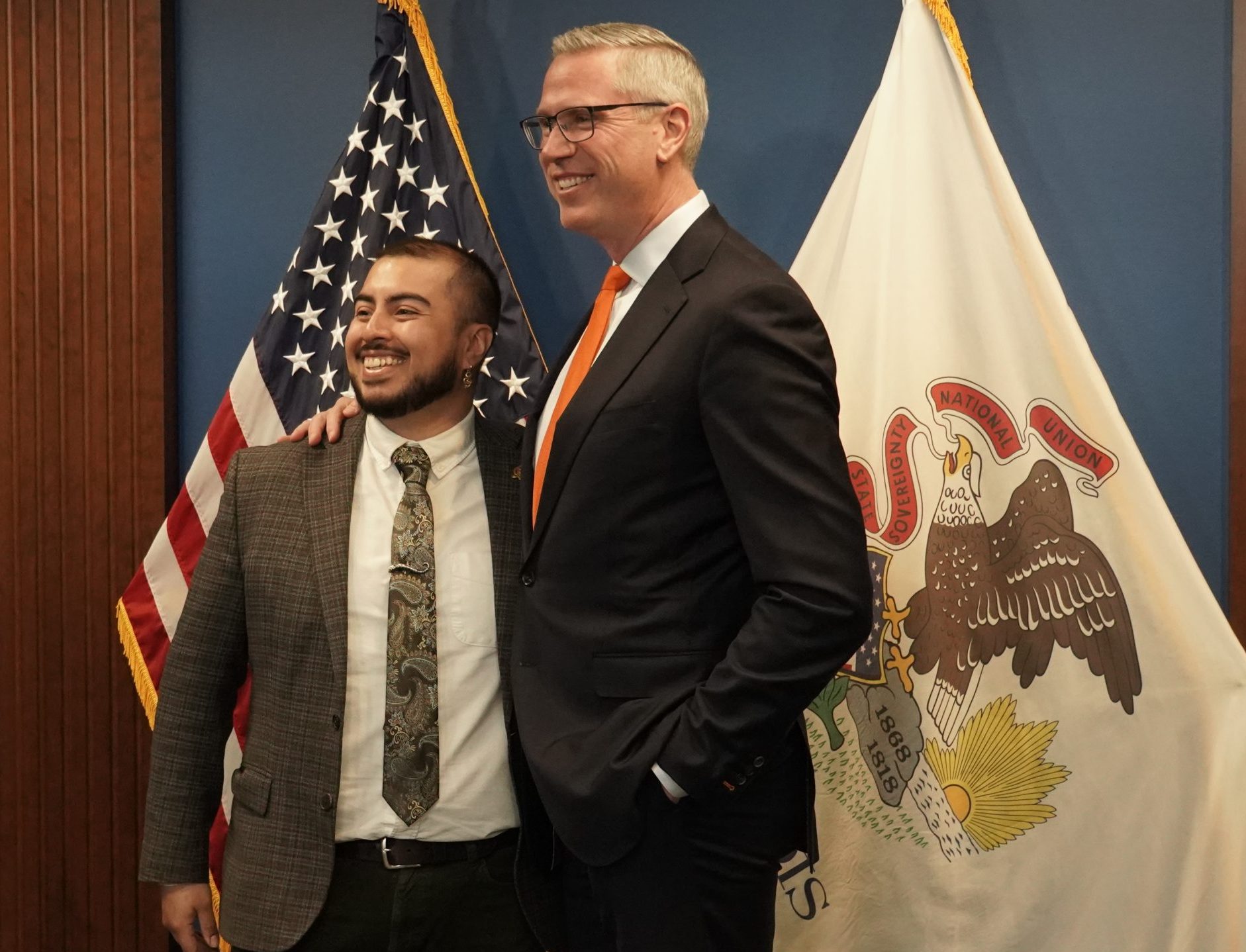 two men in suits standing in front of the illinois flag and united states flag