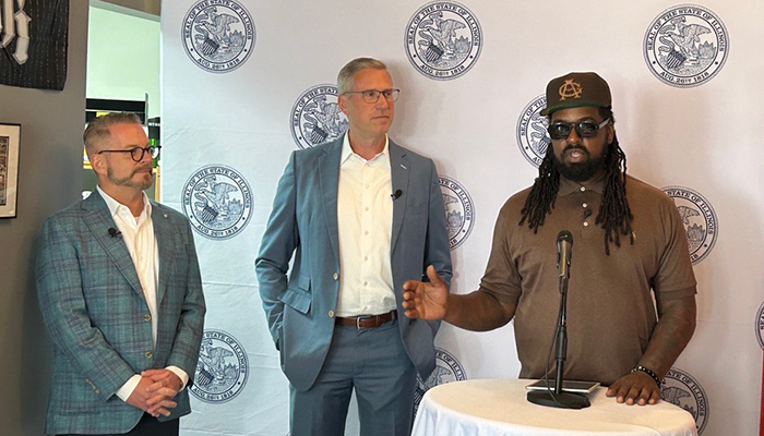 Shermann Dilla Thomas speaks at a microphone as Treasurer Frerichs and another man watch