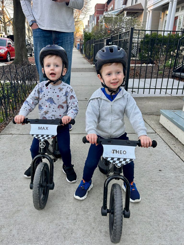 Twin toddler boys on small bicycles on a sidewalk