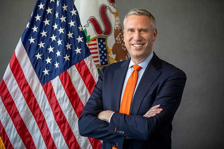 Treasurer Frerichs wearing a dark suit and orange tie, with his arm crossed. U.S. and Illinois flags are to his right.