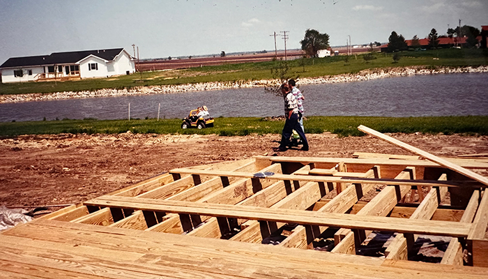 Two men walk along a dirt lot where wood framing for a house is being built