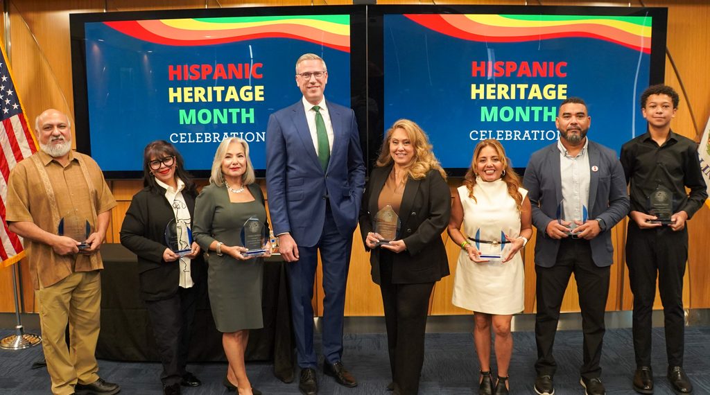 Eight people standing in front of an electronic sign that says Hispanic Heritage Month. Treasurer Frerichs stands with the four women and three men who received awards at the Heritage Month celebration.